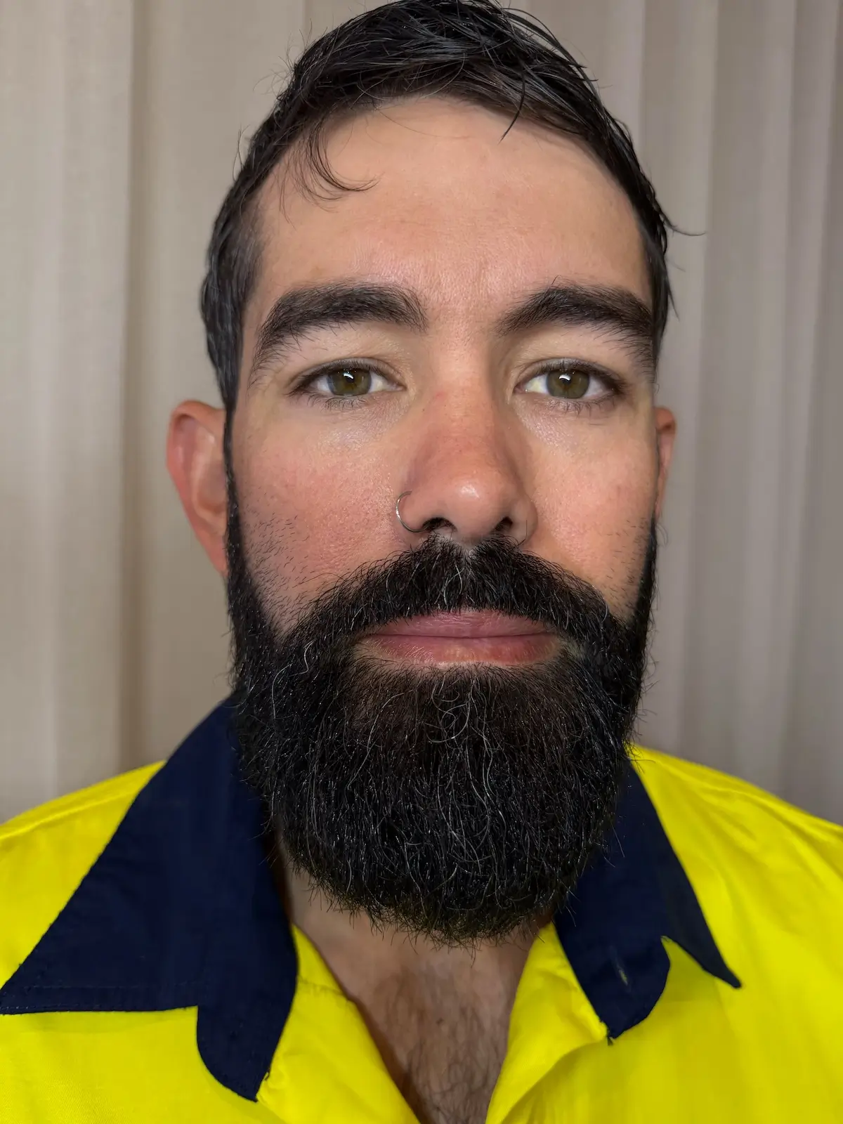 Man with a beard and nose ring wearing a high-visibility yellow shirt with dark blue collar, standing in front of a plain background.