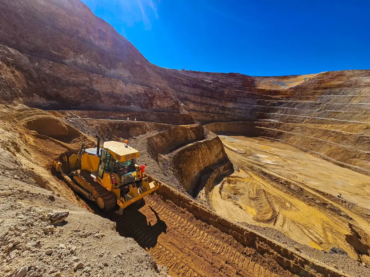A large yellow bulldozer works on the steep side of an open-pit mine. The pit has terraced layers with exposed earth and rocks under a clear blue sky. Tracks are visible, indicating recent activity.