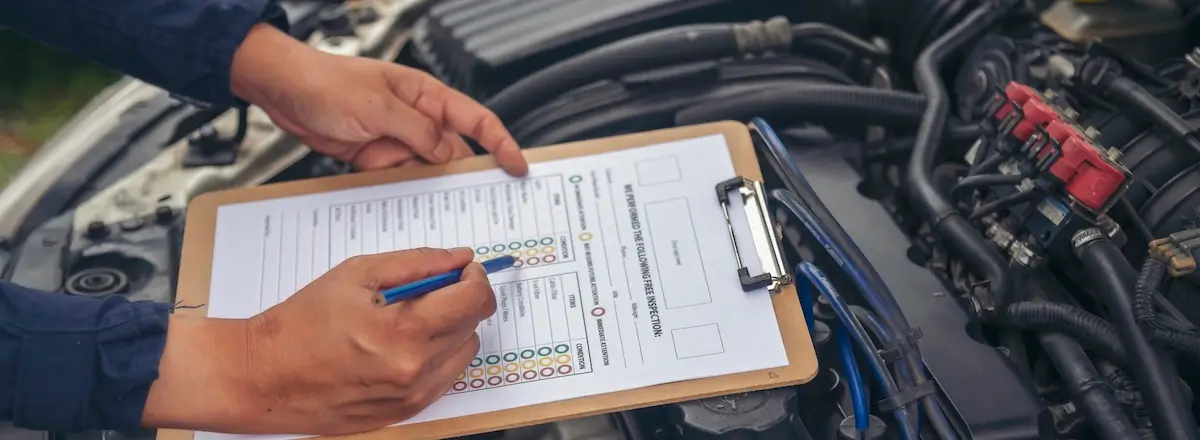 A person is holding a clipboard with a vehicle inspection checklist. They are marking boxes with a pen. The open car hood reveals engine components, suggesting an ongoing maintenance or inspection task.