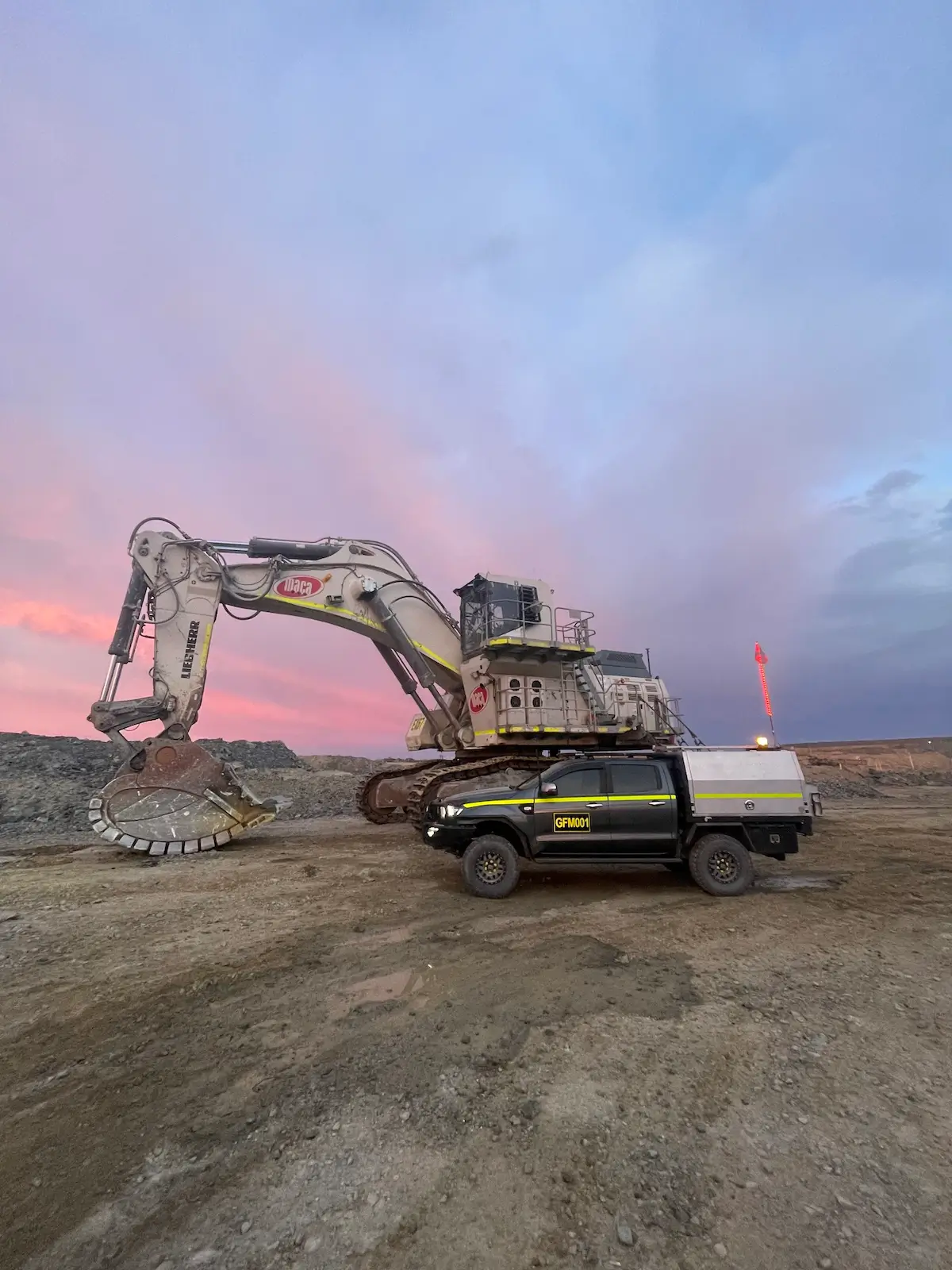 A large excavator with a massive shovel attachment is parked on a dirt site under a colorful sky at dusk. In front of it, a black and silver utility truck with the logo