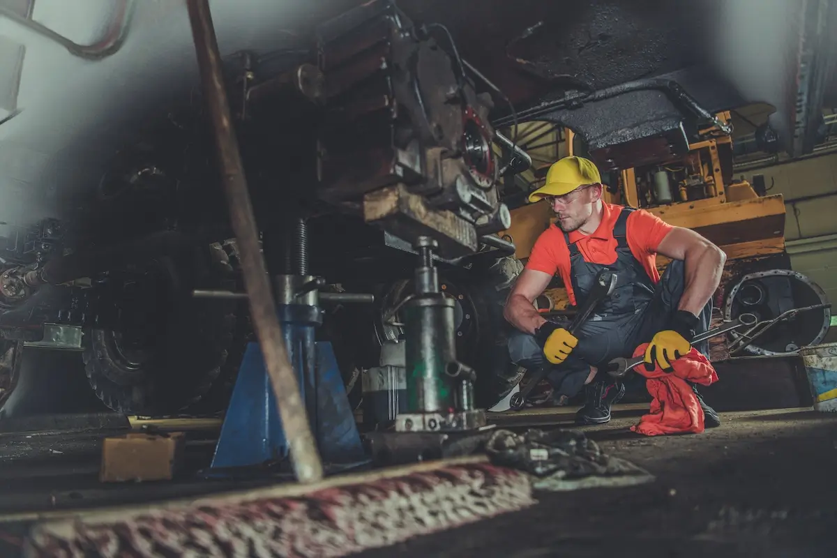 A worker in a red shirt and yellow cap crouches under a vehicle, focusing intently on a mechanical component. The dim, industrial setting highlights his concentration on the task at hand. Various tools and machinery parts are visible around him.