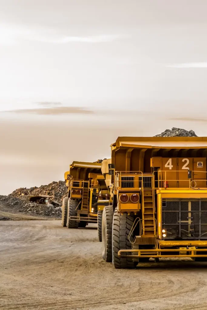 Three large yellow dump trucks are lined up on a dirt road at a mining site, with mounds of earth and rock in the background under a cloudy sky.