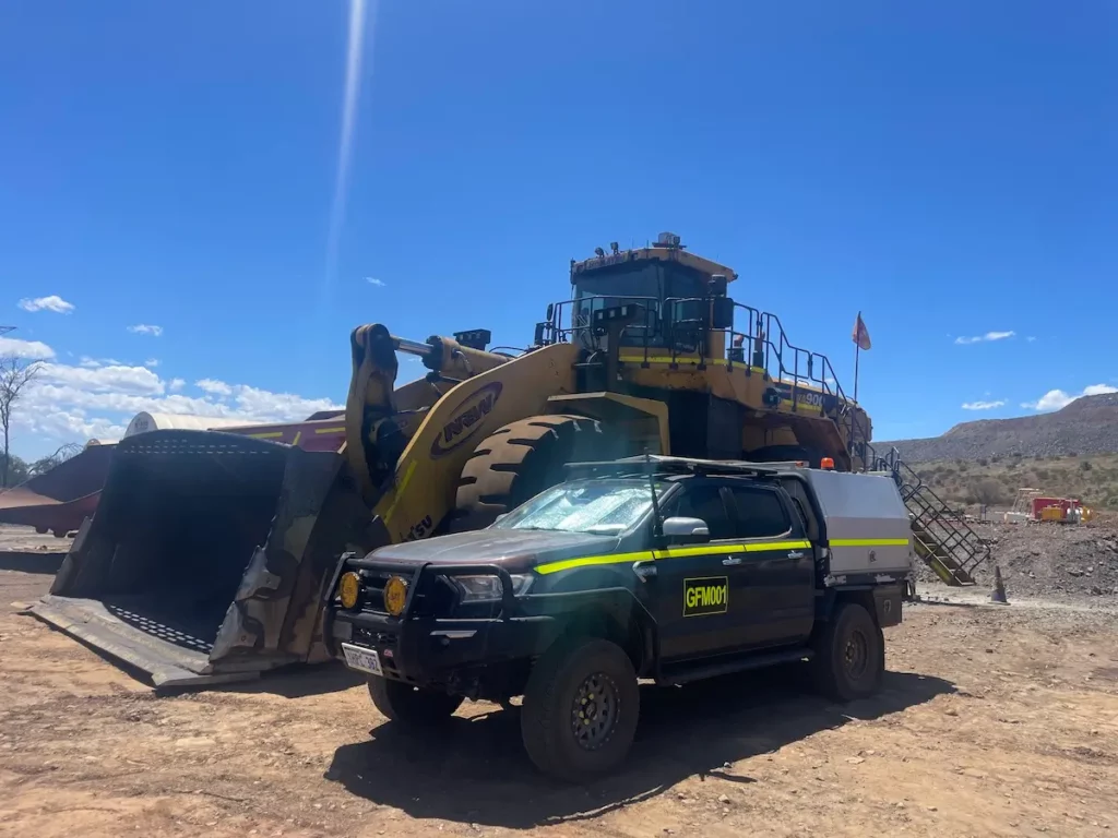 A black utility vehicle with yellow stripes and "GFAMM" on the side is parked on a dirt lot in front of a large yellow industrial loader. The sky is clear and blue, with hills visible in the background.