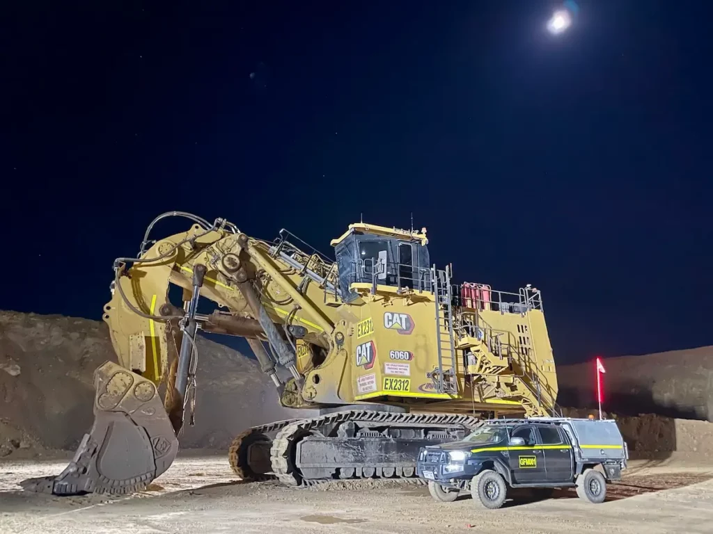 A large excavator is parked on a construction site at night under a bright moon. A smaller utility vehicle is parked beside it, showcasing the size difference. The sky is clear and dark, highlighting the machinery and the illuminated work area.