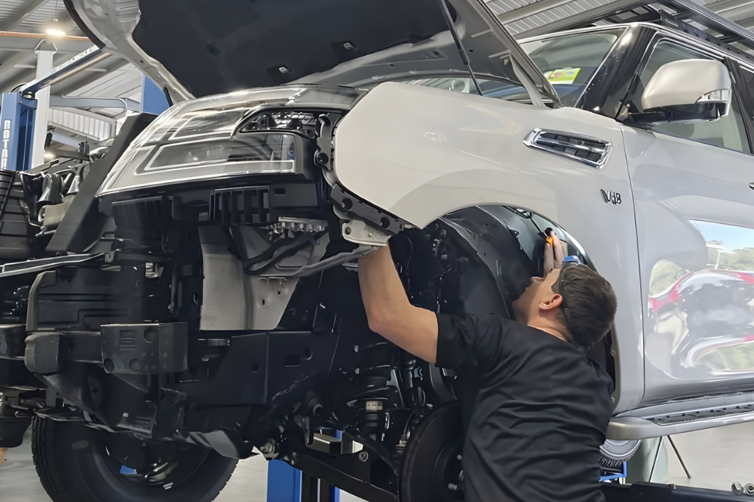 A mechanic works under the front wheel well of a white SUV with its hood open and bumper removed inside an auto repair shop.