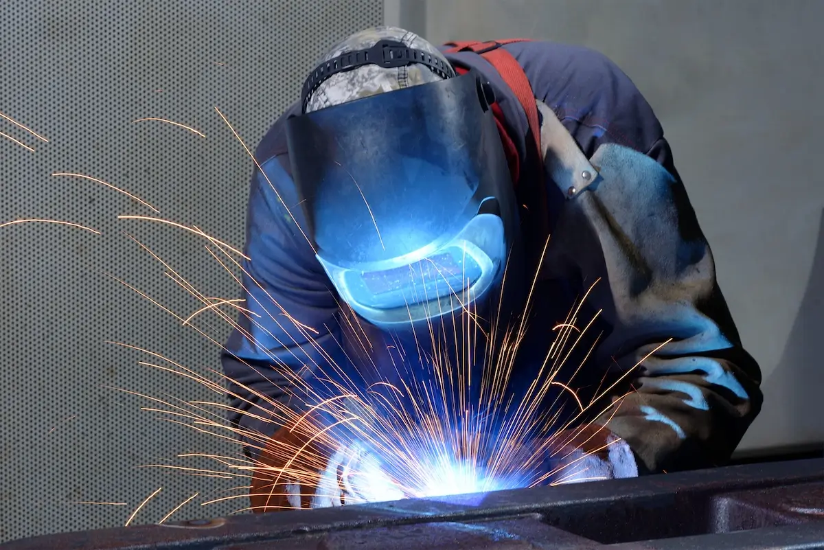 A welder wearing protective gear, including a helmet and gloves, works on a metal piece. Bright sparks fly outward as he joins the metal, highlighting the intense glow from the welding process. The background is a muted indoor setting.