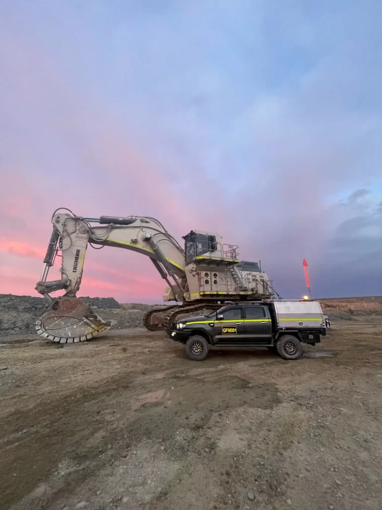 A large excavator with a wide bucket is parked on a dusty construction site under a pink and blue sky. In front of it is a black utility vehicle with a silver canopy, displaying the word "Orix" on the side.