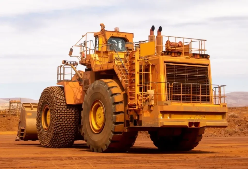 A large, heavy-duty mining truck with massive wheels and a rugged design is parked on a dirt surface. The truck is primarily yellow with some visible wear and marks. A partly cloudy sky is in the background.