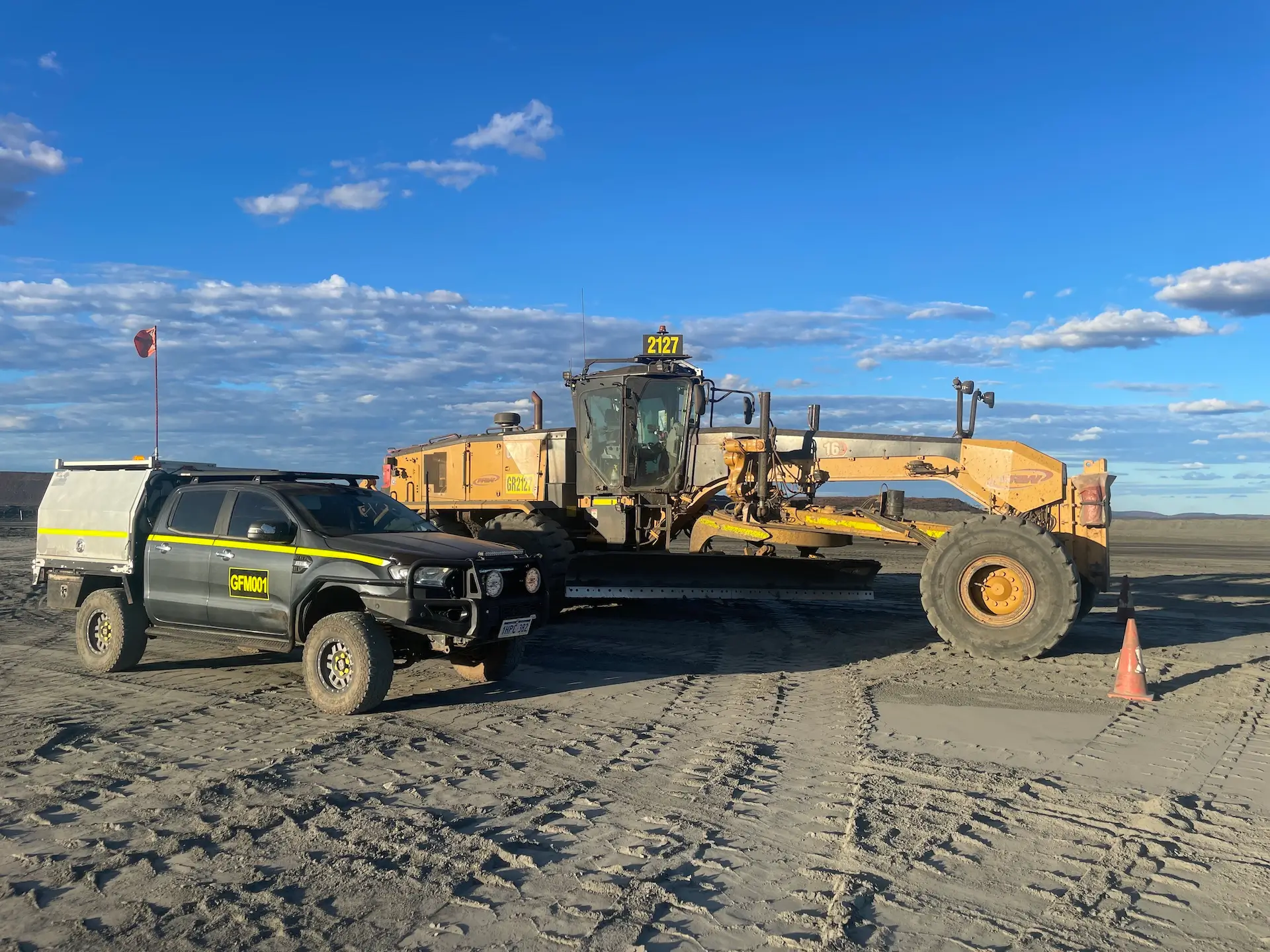 A large yellow grader and a black utility vehicle with a yellow logo are parked on a sandy construction site under a blue sky with scattered clouds. An orange traffic cone is nearby.