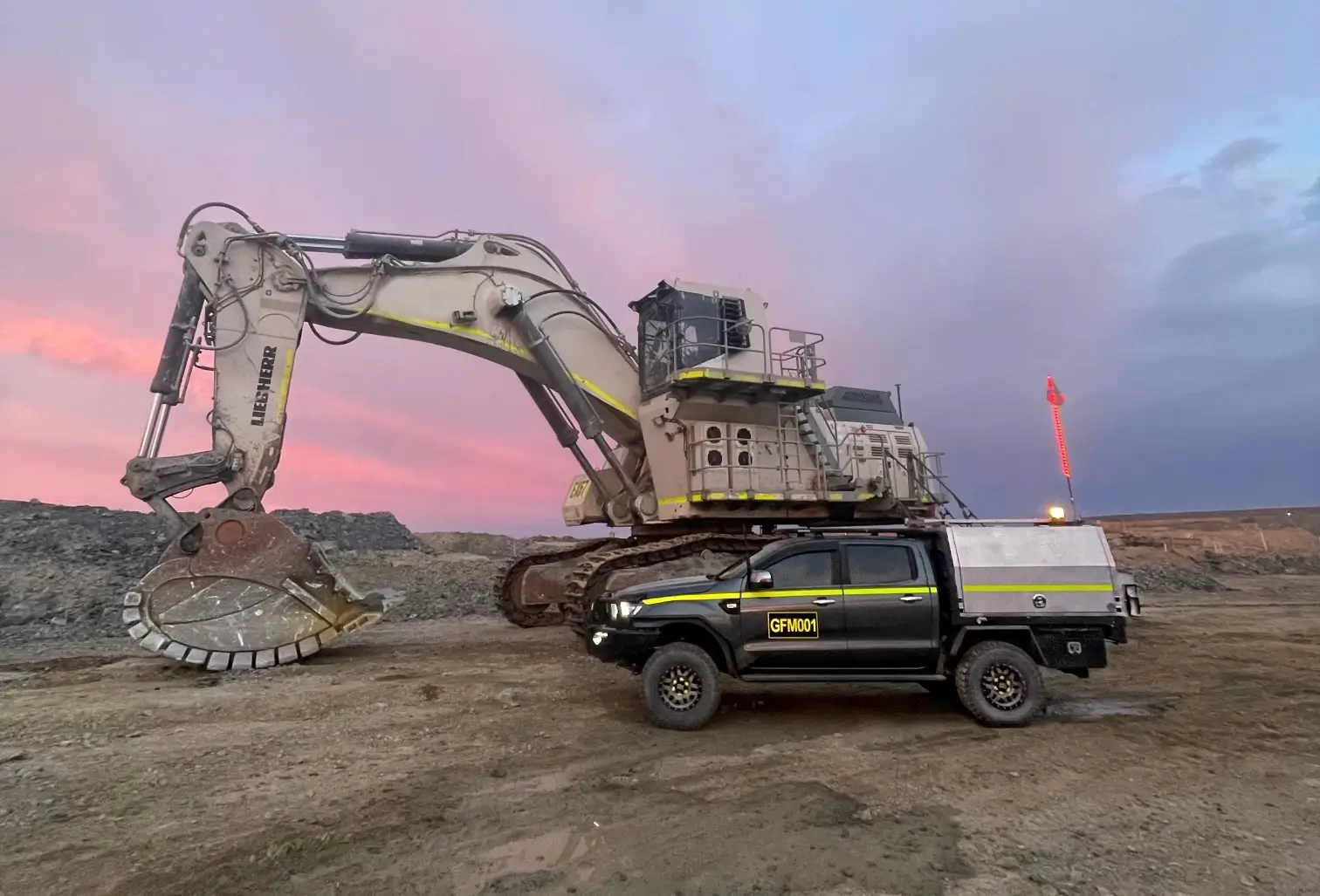 A large excavator and a rugged utility vehicle are parked on a construction site during sunset. The sky is a blend of pink and purple hues, and the ground is earthy and bare. The environment suggests ongoing industrial work.