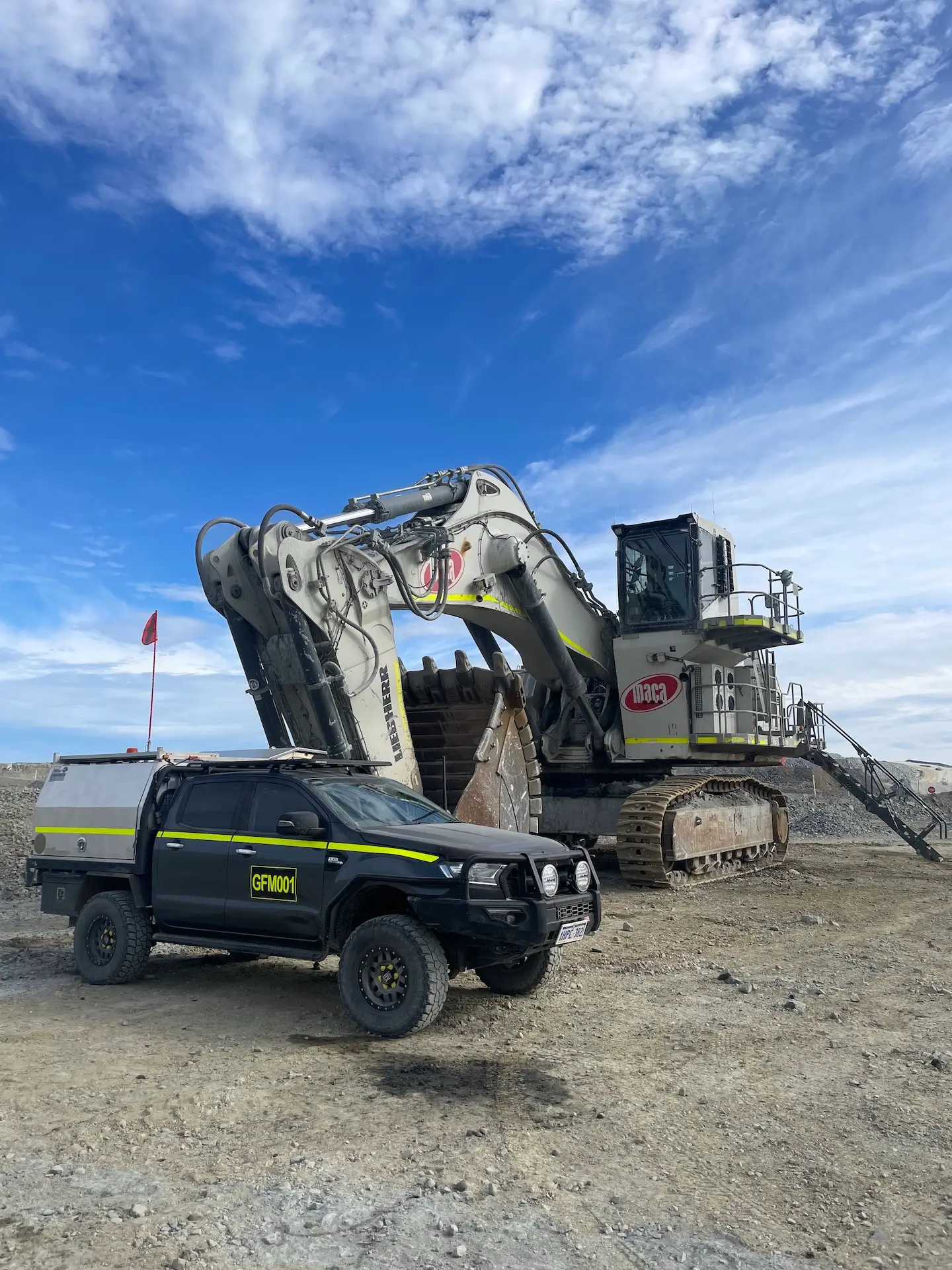 A large excavator with a shovel arm is parked on a construction site under a blue sky with clouds. In front of the excavator, there's a black and yellow utility vehicle with a canopy and visible license plate. A red flag is in the background.