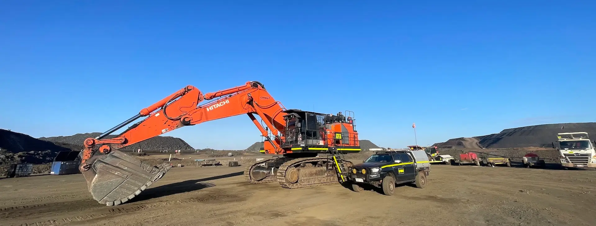 An orange construction excavator is parked on a dirt site under a clear blue sky. Nearby, a dark-colored utility vehicle and several trucks are present. Piles of dirt and distant hills are visible in the background.