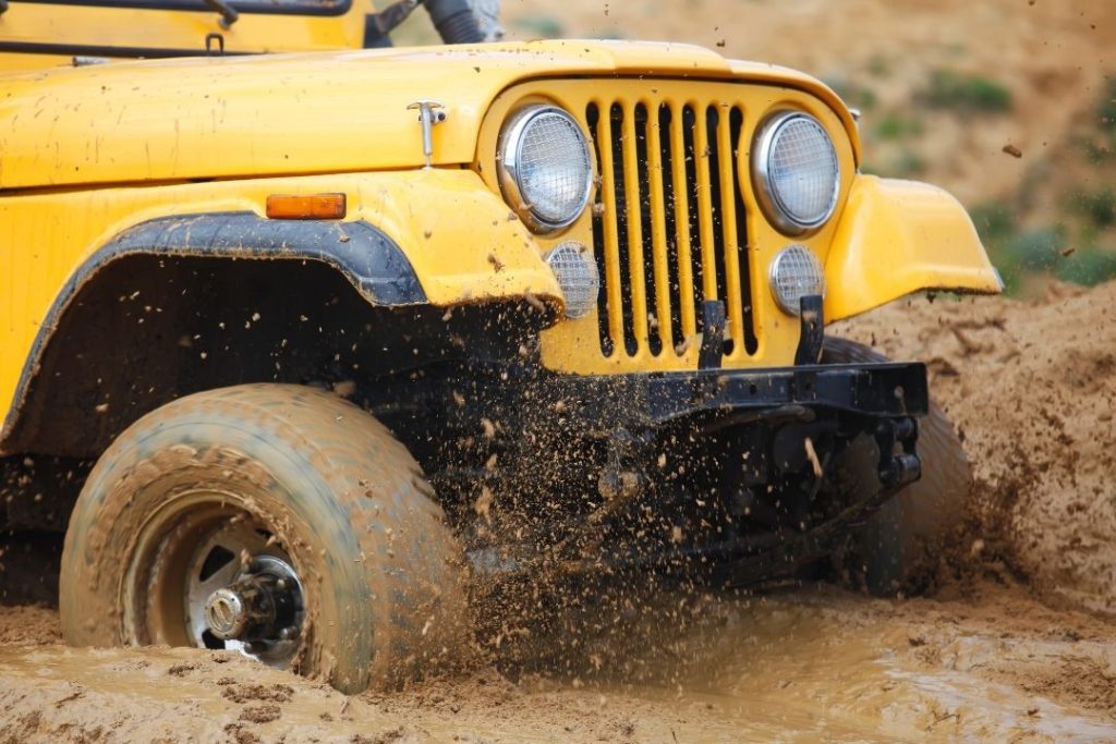 Close-up of a yellow off-road vehicle driving through muddy terrain, mud splashing around its spinning front tires—a perfect scene to highlight the importance of mechanical maintenance and repair for peak performance.
