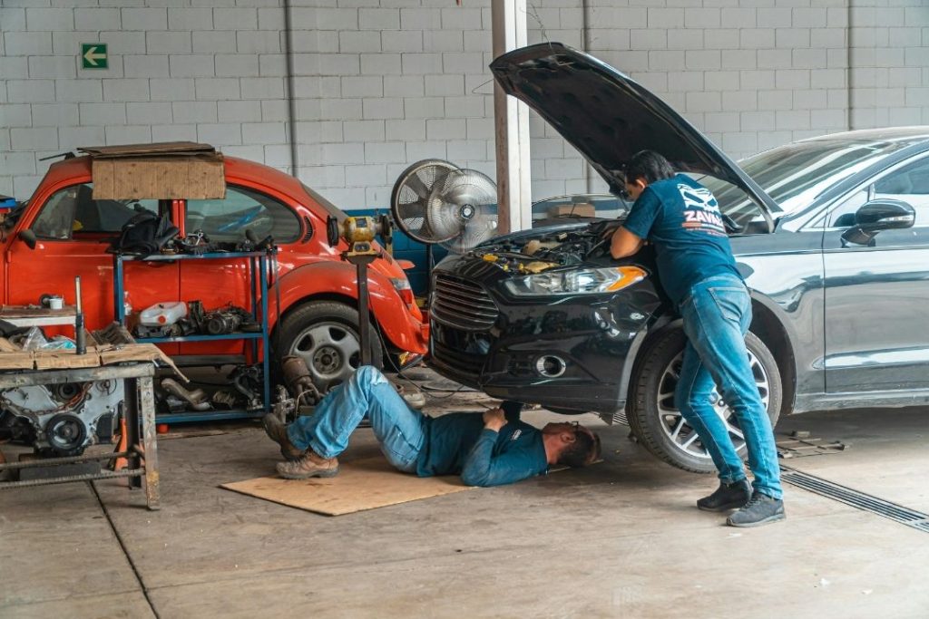 Two mechanics work on a black car in an auto repair shop, specializing in mining and civil mechanical services; one is under the car on a mat, while the other leans over the engine bay. A red vintage car and various tools are visible in the background.