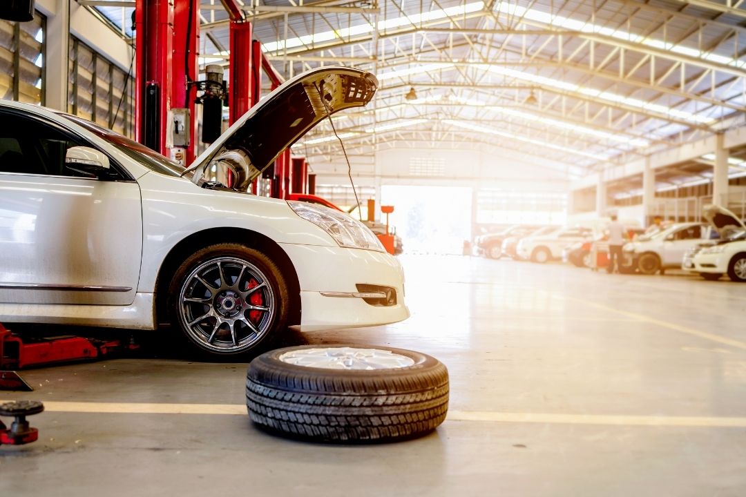 A white car with its hood open is lifted on a hydraulic jack in a spacious auto repair shop specializing in mining and civil mechanical services. A wheel lies on the floor in the foreground, with other cars parked in the background.