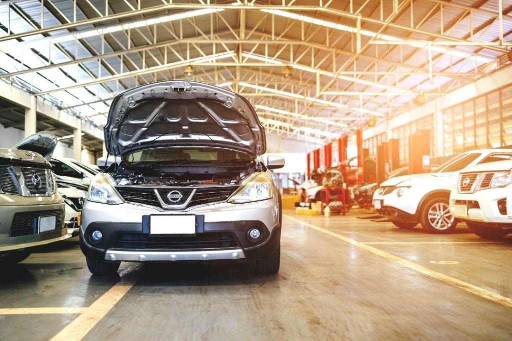 A silver Nissan car with its hood open is parked inside a large, brightly lit auto repair shop offering diesel mechanical services, surrounded by other vehicles and workstations. Sunlight streams through the windows above.