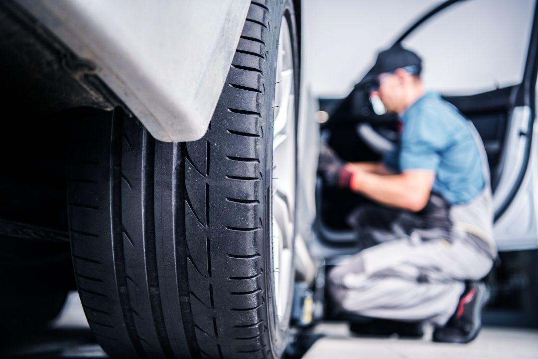 Close-up of a car tire with a mechanic in uniform and cap kneeling beside an open car door in the background, performing maintenance or inspection work. The focus is on the tire tread.