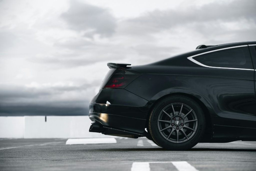 The rear side of a sleek black sports car parked on a rooftop with overcast skies in the background, highlighting its taillights, rear wheel, and spoiler—primed for mechanical maintenance and repair.