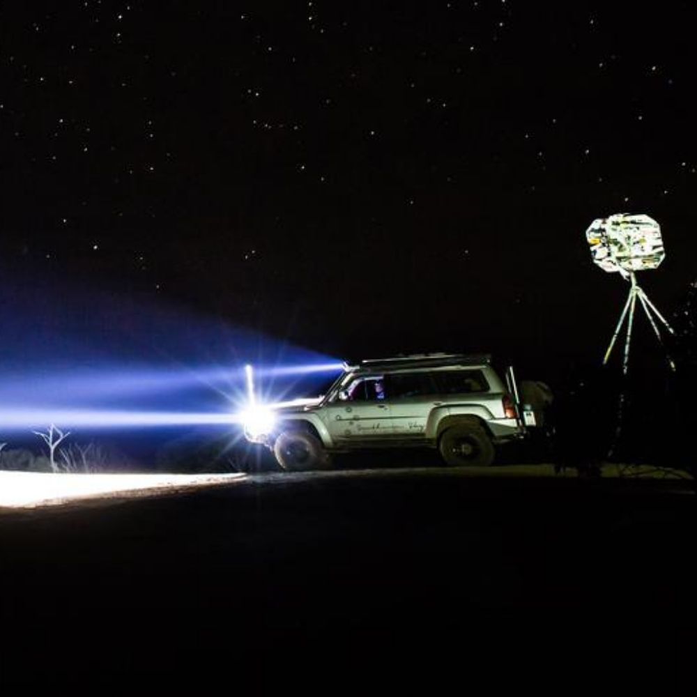 A parked off-road vehicle shines bright headlights into the night, illuminating the dark landscape under a star-filled sky. A tripod with a covered object stands nearby.