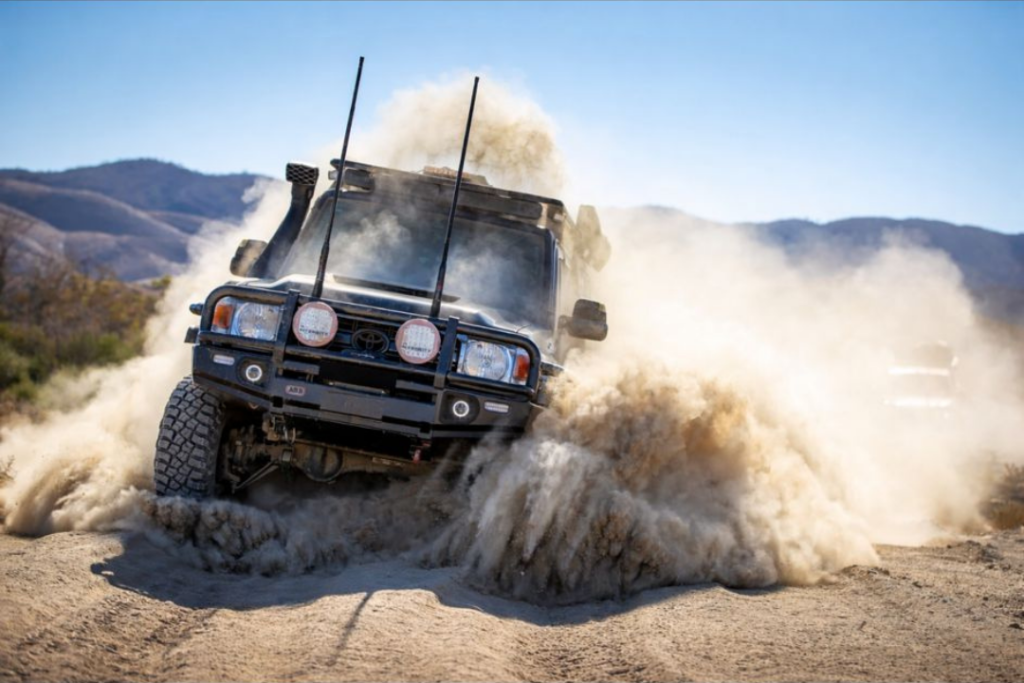 A black off-road SUV kicks up a large cloud of dust while driving over rough terrain—ideal for mining and civil mechanical services—with mountains and a clear blue sky in the background.