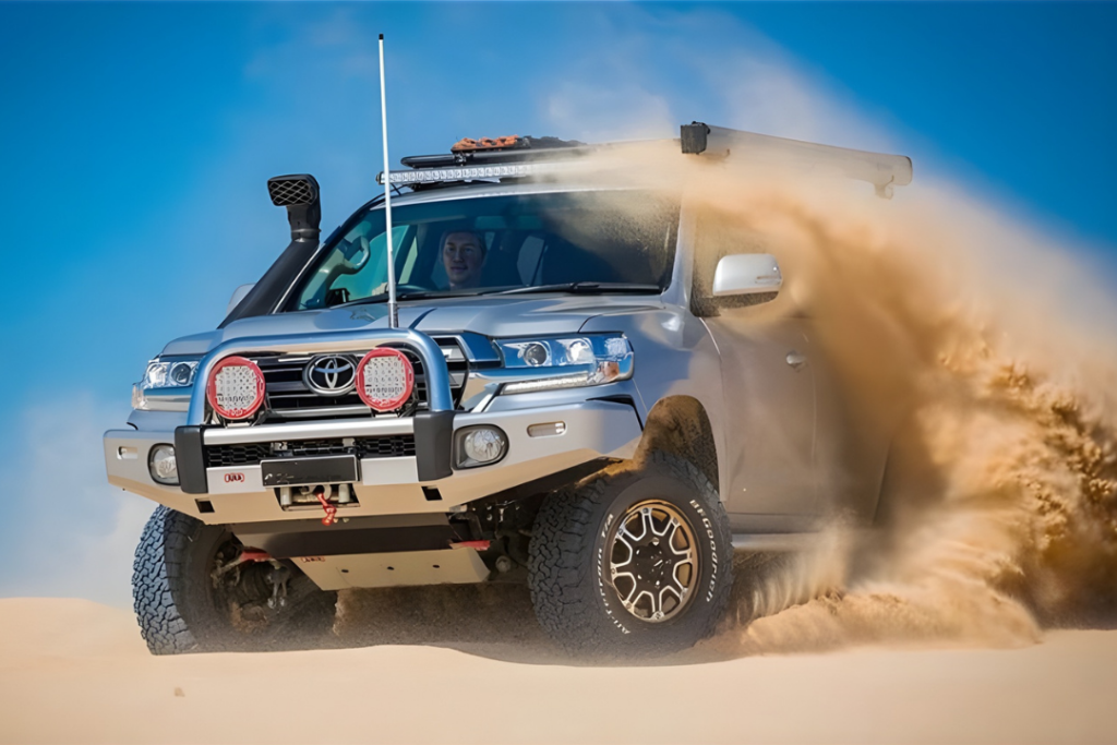 A silver off-road vehicle, used in mining and civil mechanical services, drives quickly through sand dunes, kicking up a large cloud of sand around its tires under a clear blue sky.