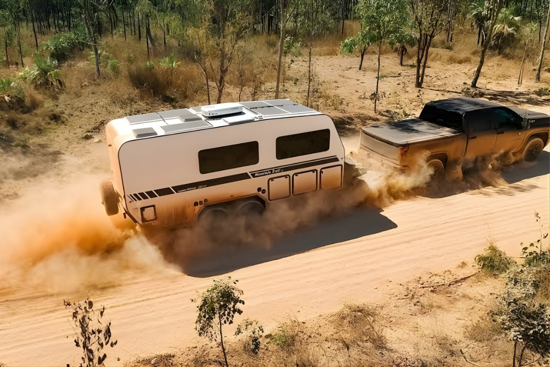 A pickup truck, well-kept through mechanical maintenance and repair, tows a white travel trailer, stirring up dust while driving on a dirt road surrounded by trees and dry vegetation.