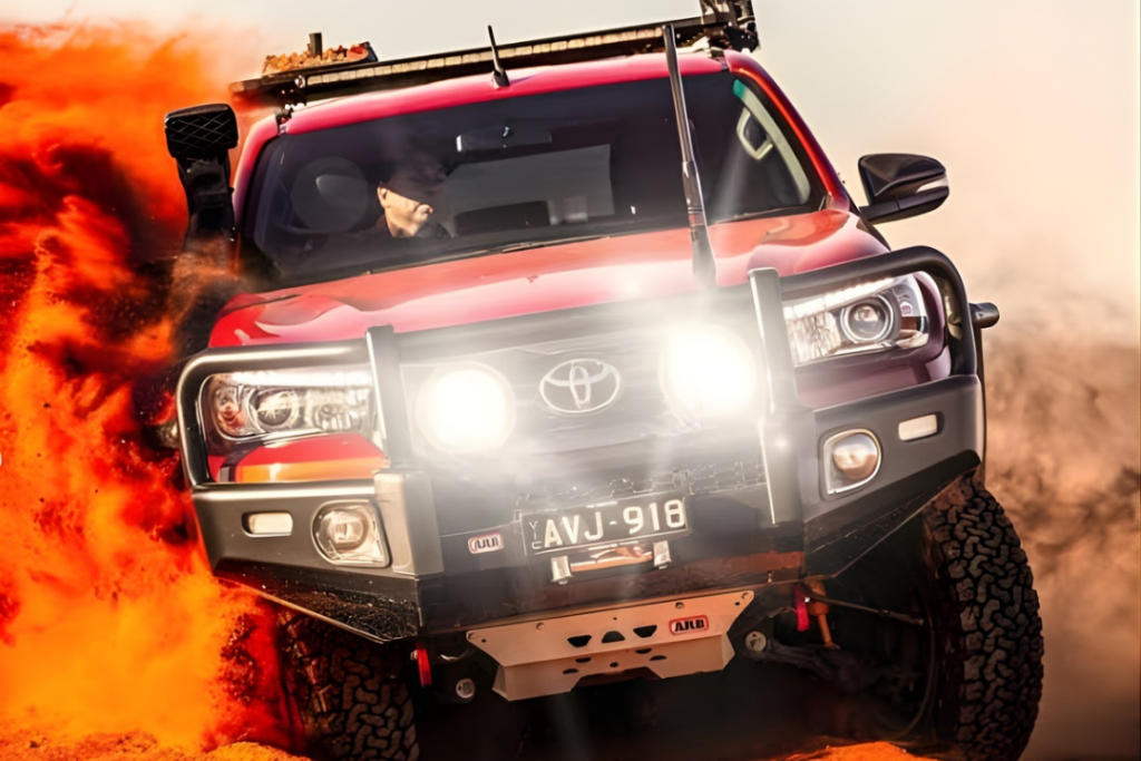 A red Toyota truck, outfitted for mining and civil mechanical services, drives through orange sand with headlights on, kicking up a large spray of dust. The vehicle boasts off-road accessories and a visible license plate.