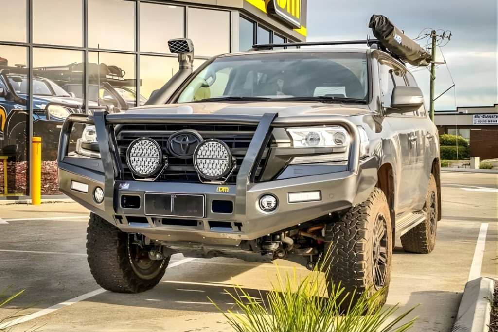 A grey Toyota SUV equipped with a bull bar, spotlights, roof rack, and snorkel advertises diesel mechanical services as it’s parked in a lot near a building with large windows and yellow signage.